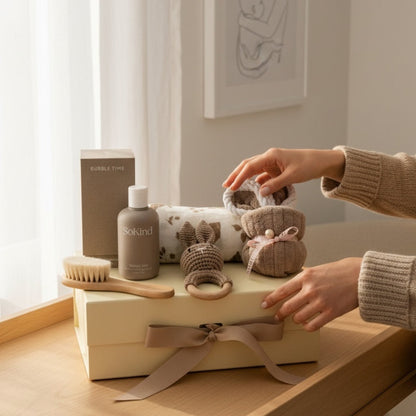 Hands arranging a neutral baby gift set on a gift box, featuring organic baby wash, wooden brush, crochet bunny rattle, muslin cloth and soft towel in a natural light setting