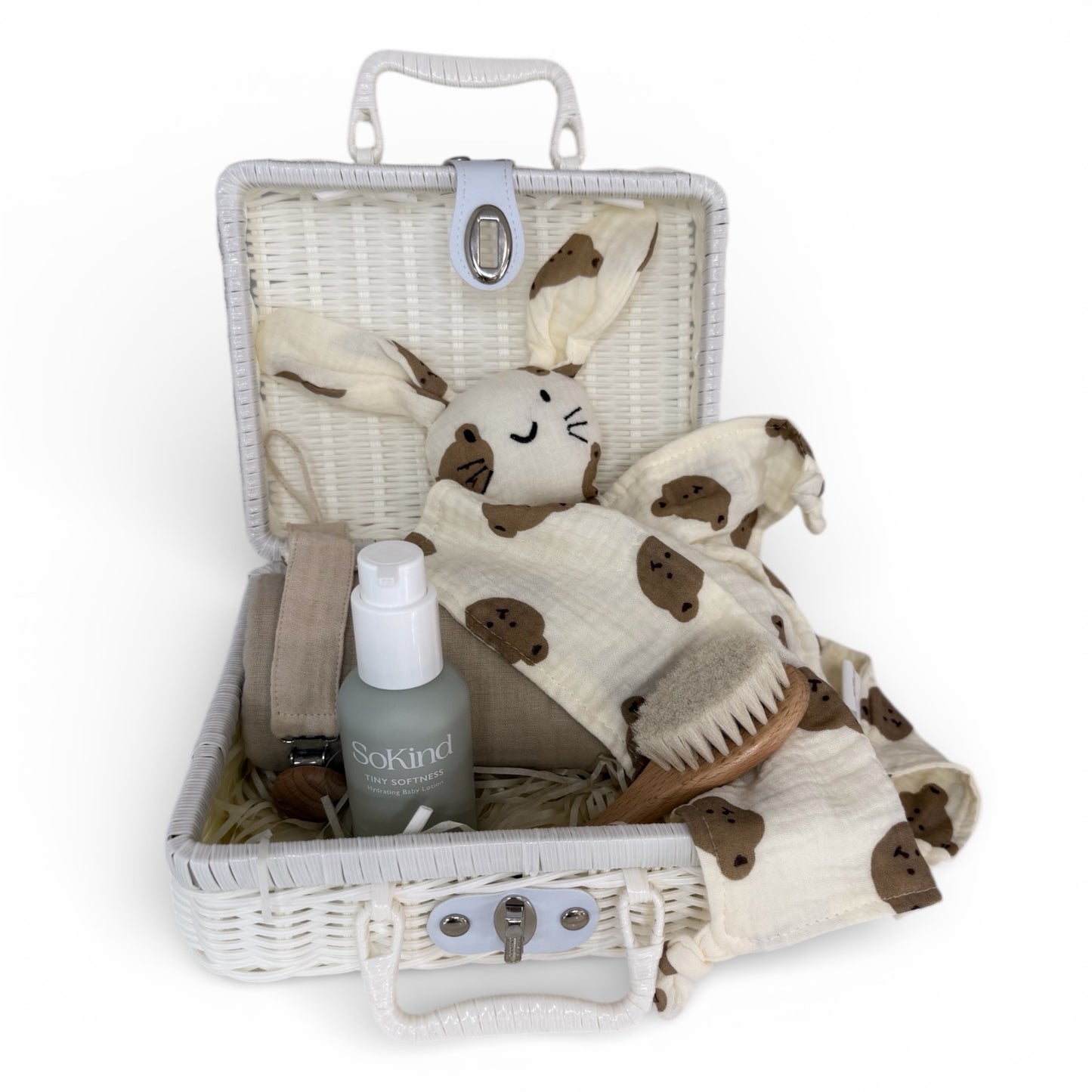 White wicker baby gift suitcase open with beige muslin cotton, a bunny comfort, baby moisturiser bottle, wooden baby brush and pacifier holder styled on a white background 