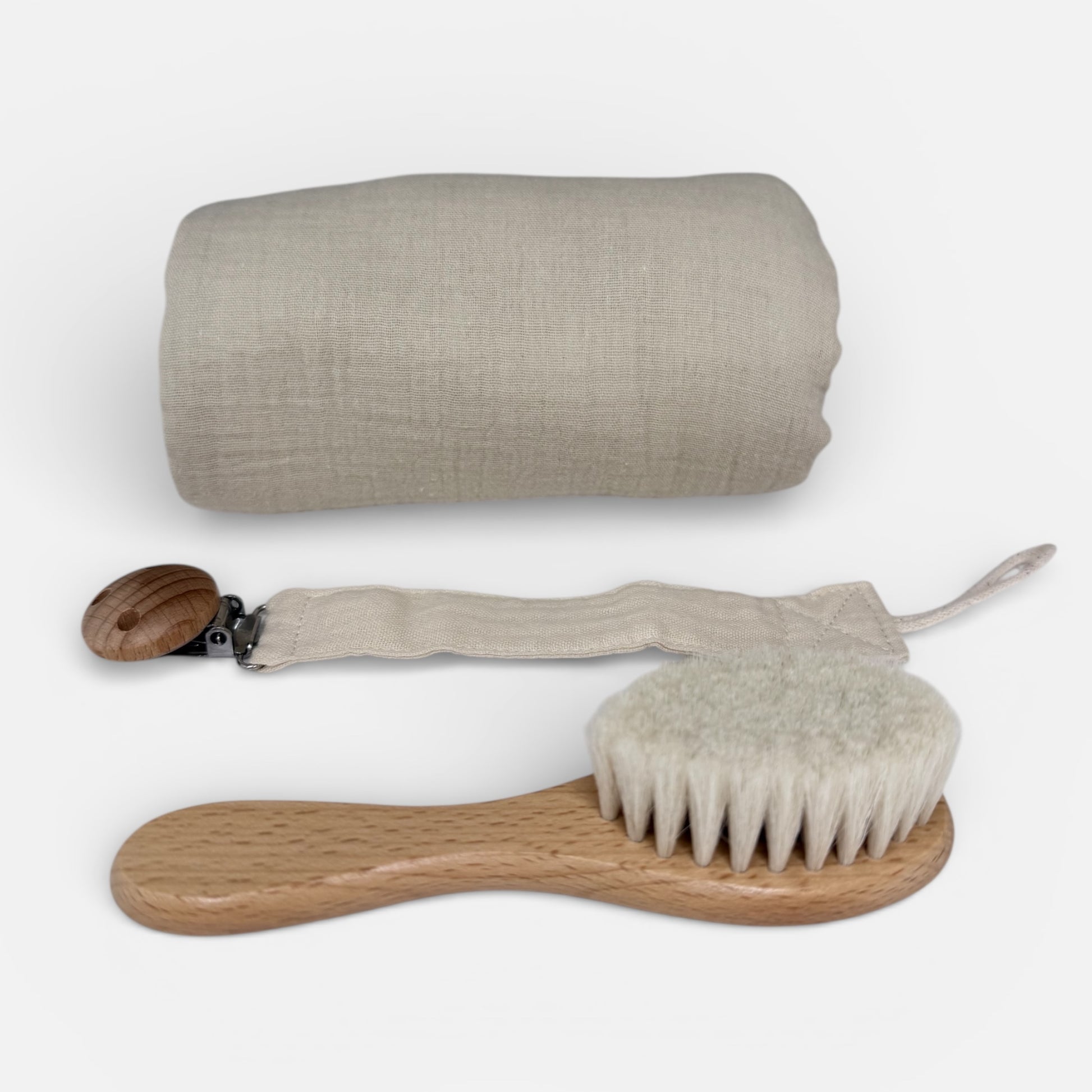 Beige muslin baby cloth rolled neatly, beige pacifier holder with wooden clip and natural wooden baby brush displayed on a white background 