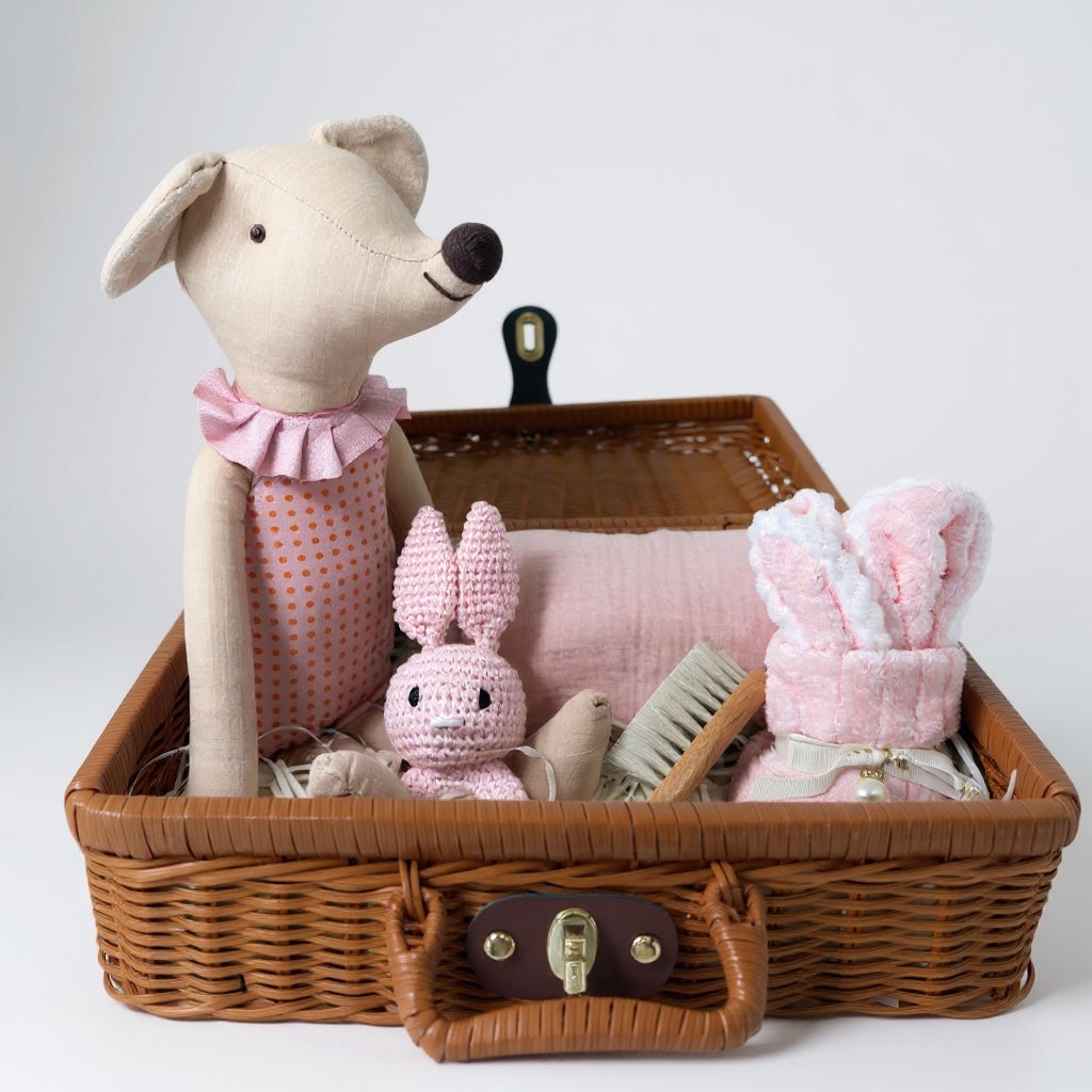 Pink baby gift hamper arranged in a wicker suitcase with soft toys, muslin cloth, baby face cloth and wooden brush on white background 