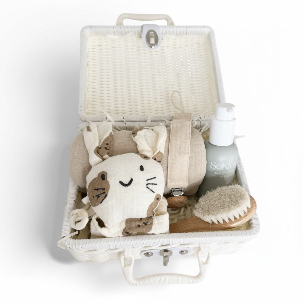 Open white wicker baby gift suitcase with a beige baby comfort blanket, muslin cloth, baby moisture and pacifier holder arranged neatly inside on a white background