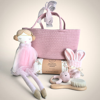 Blush baby gift hamper with wooden pink basket, organic cotton muslin swaddle, soft doll, bunny rattle, bunny towel and wooden brush on neutral background