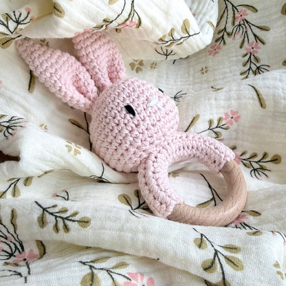 Crochet bunny rattle with wooden ring placed on softly wrinkled muslin blanket, neutral textured background, baby gift detail.