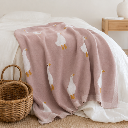 Pink knitted organic cotton baby blanket with white goose pattern draped over cot beside woven basket in nursery setting