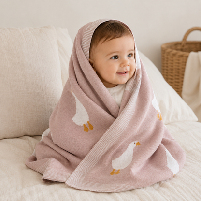 Smiling baby wrapped in pink knitted organic cotton baby blanket with white goose pattern sitting on bed by Wrap With Love