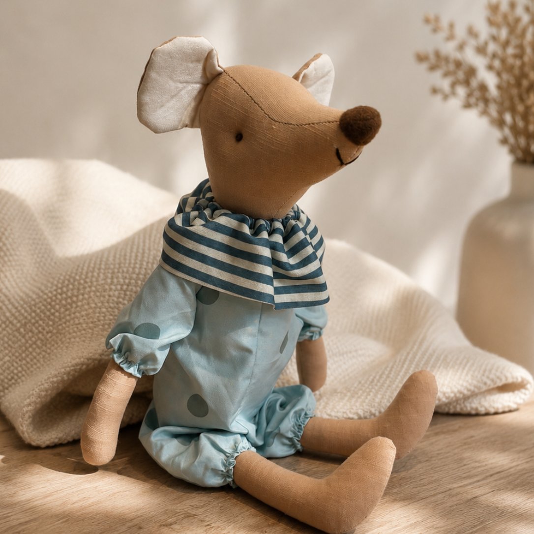 Soft mouse plush toy wearing a sage outfit sitting on a neutral knitted blanket in a boutique nursery setting
