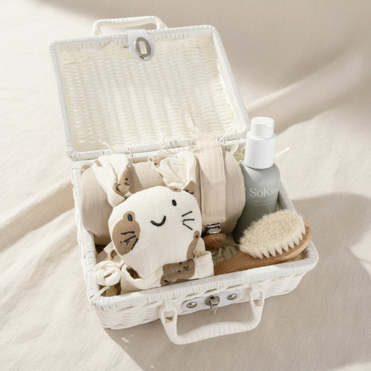 Neutral baby gift hamper in a white wicker suitcase with baby comfort blanket, muslin cloth, baby brush and skincare essentials on a soft beige background 