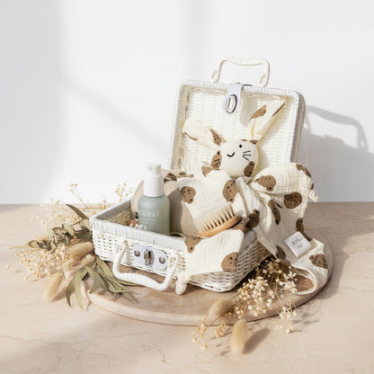 Neutral bunny baby gift hamper in a white wicker suitcase with muslin swaddle, comfort toy, baby brush and skincare essentials on a soft beige background 