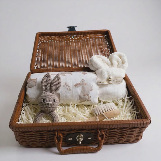 Ivory baby gift hamper with wicker suitcase, including muslin swaddle, bunny rattle, rolled baby face cloth and wooden brush on white background