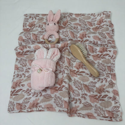 Baby gift essentials including pink muslin swaddle, crochet bunny rattle, baby brush and bunny face washer, laid out together.