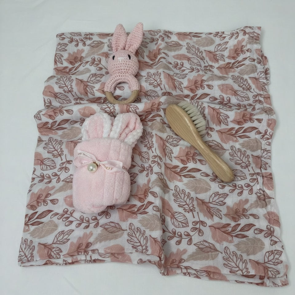Baby gift essentials including pink muslin swaddle, crochet bunny rattle, baby brush and bunny face washer, laid out together.